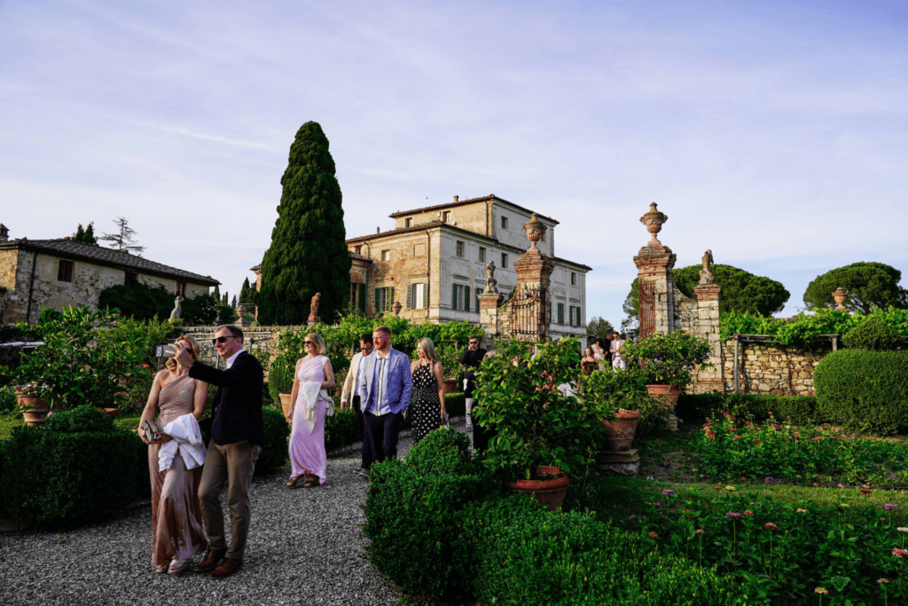 Guests in formal attire walk through a lush garden with flowers and greenery, in front of a large historic villa under a clear sky, during what appears to be a festive event, as part of an exclusive incentive travel programme.
