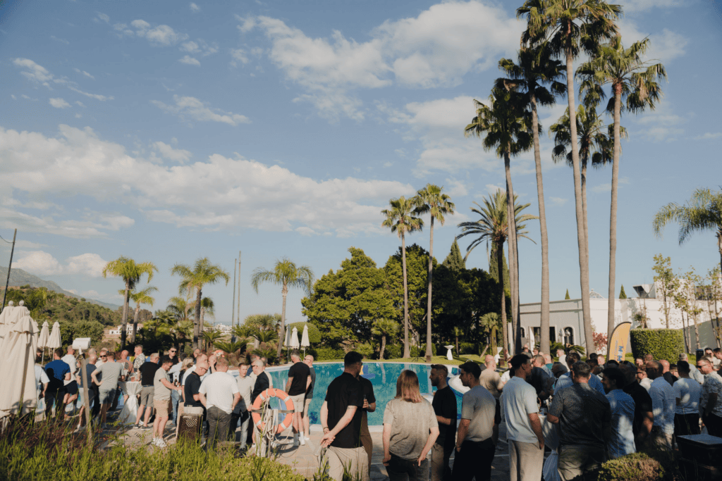 A large group of people gathers outdoors near a swimming pool, surrounded by palm trees and greenery, under a blue sky with scattered clouds, enjoying the vibrant atmosphere of an incentive travel programme.