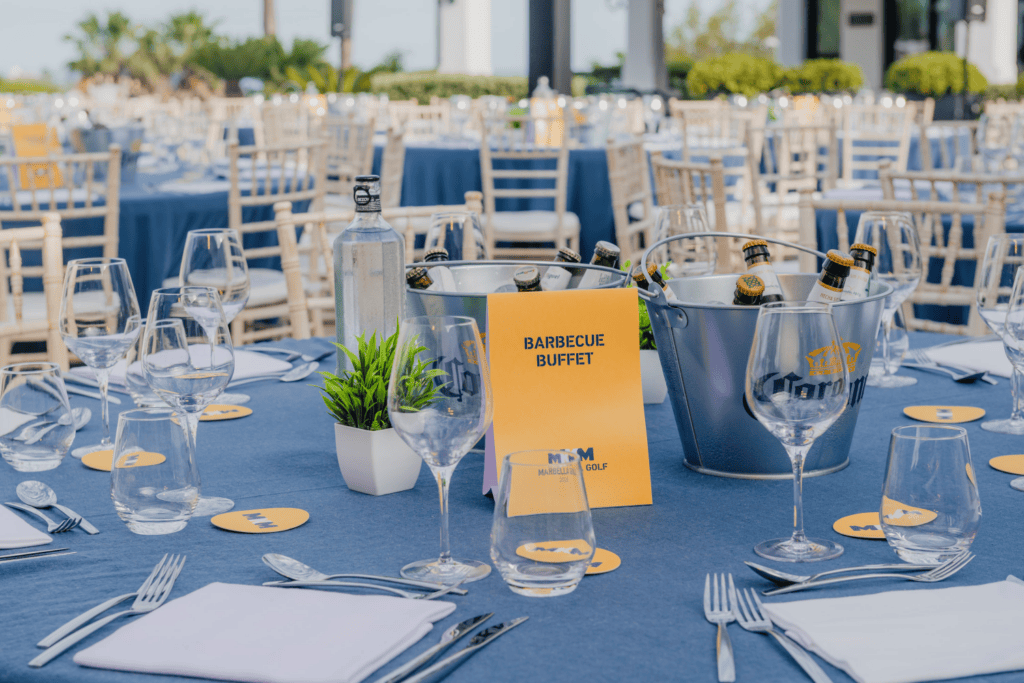 Outdoor table set for an event with blue tablecloths, wine glasses, water bottles, beer in an ice bucket, and a yellow Barbecue Buffet sign—an inviting setup perfect for your next incentive travel programme. White chairs and more tables are visible in the background.
