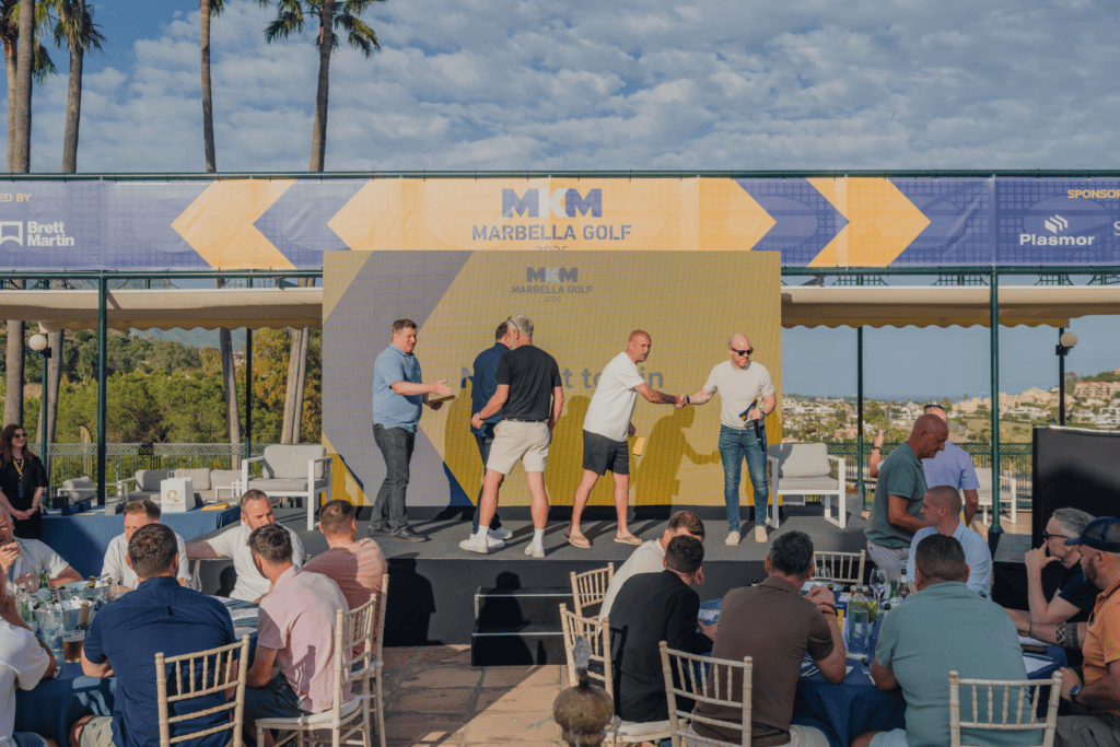 Four men stand on an outdoor stage at a Marbella Golf event, shaking hands and exchanging items as part of an incentive travel programme, with people seated at tables in the foreground and palm trees in the background.