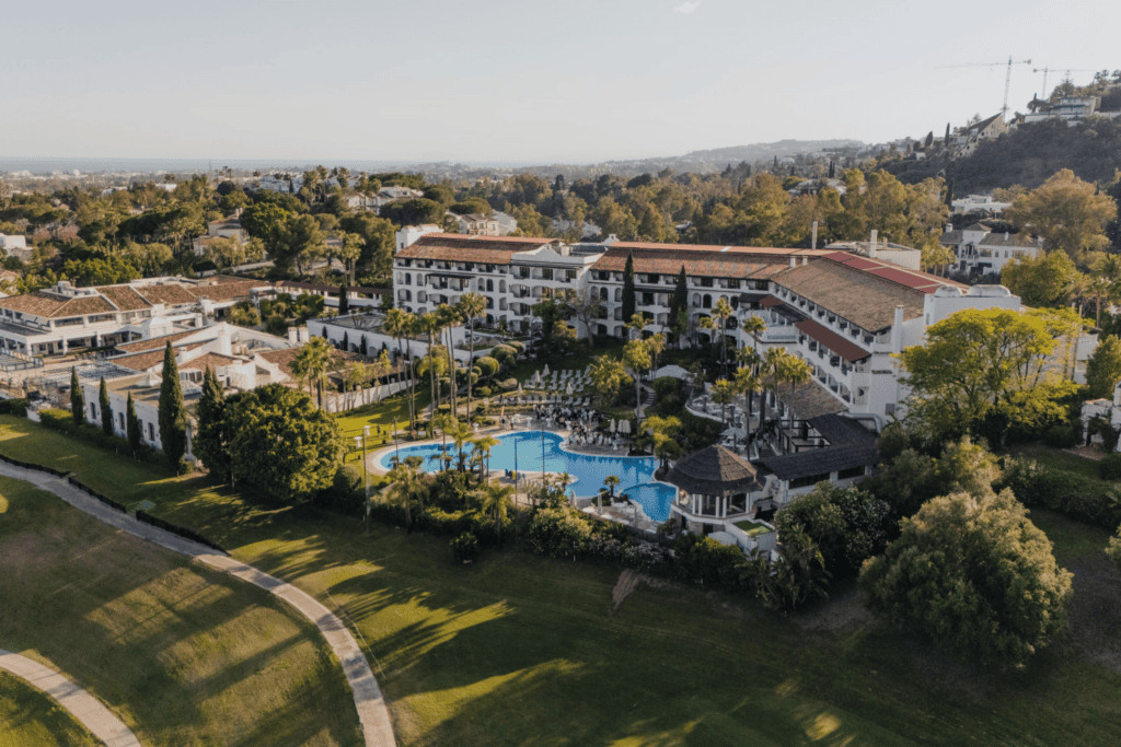 Aerial view of a resort with a large outdoor pool, surrounded by palm trees, gardens, and buildings, set near a golf course with green hills in the background—an ideal destination for an unforgettable incentive travel programme.