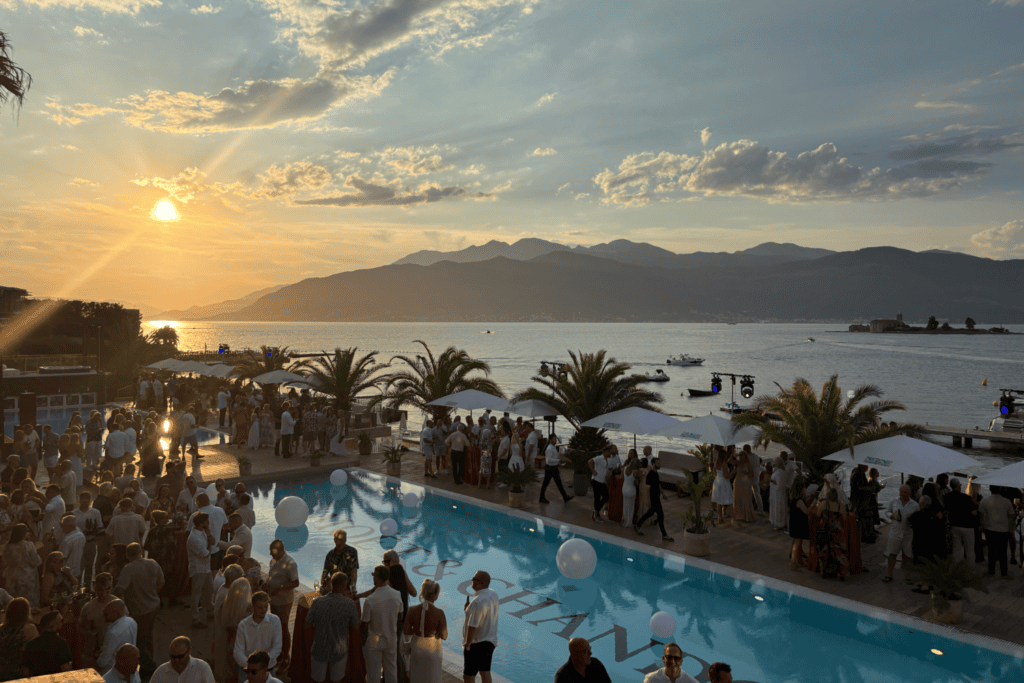 A lively crowd gathered around a pool at sunset by the sea, as part of an exclusive incentive travel programme, with palm trees swaying, mountains in the background, and boats gliding on the water—all coming together to create a truly festive atmosphere.
