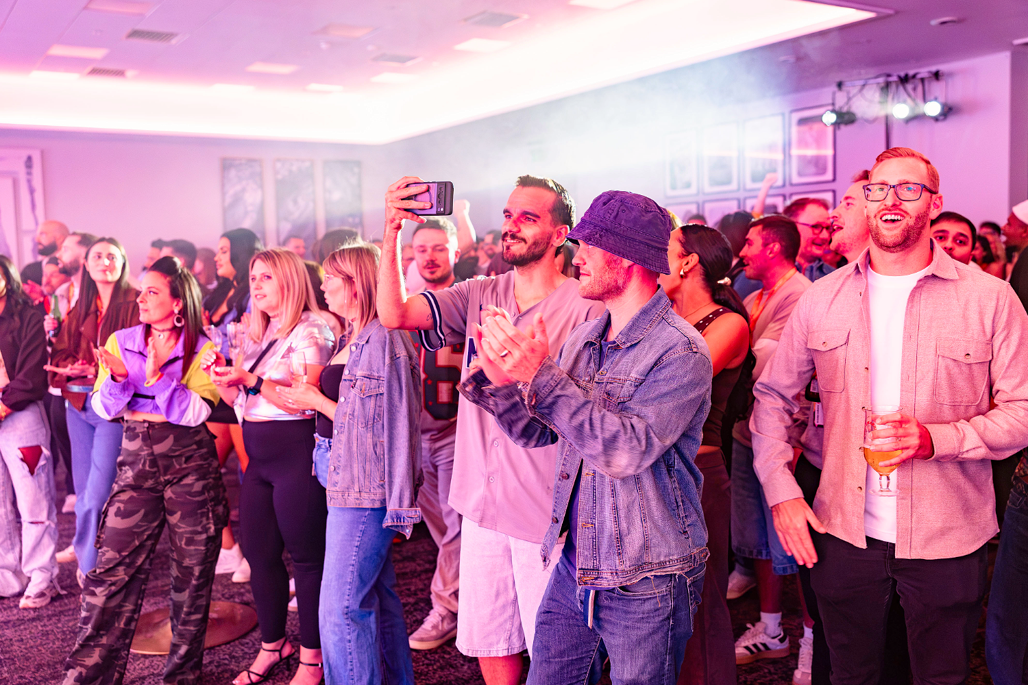 A group of people at an indoor event stand close together, some applauding, smiling, or recording with mobiles. The lighting is pink and purple, and several people hold drinks. The atmosphere appears lively and social, reflecting high event engagement among the attendees.