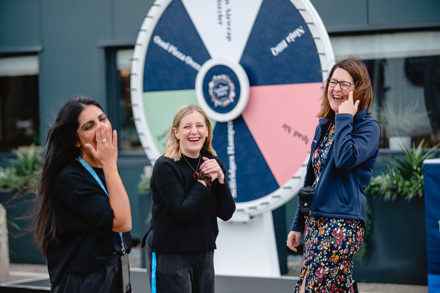 Three women laugh together in front of a large spinning prize wheel outdoors, creating memorable event experiences. Two have dark and blonde hair and wear black, while the third wears glasses and a blue jacket over a floral dress.
