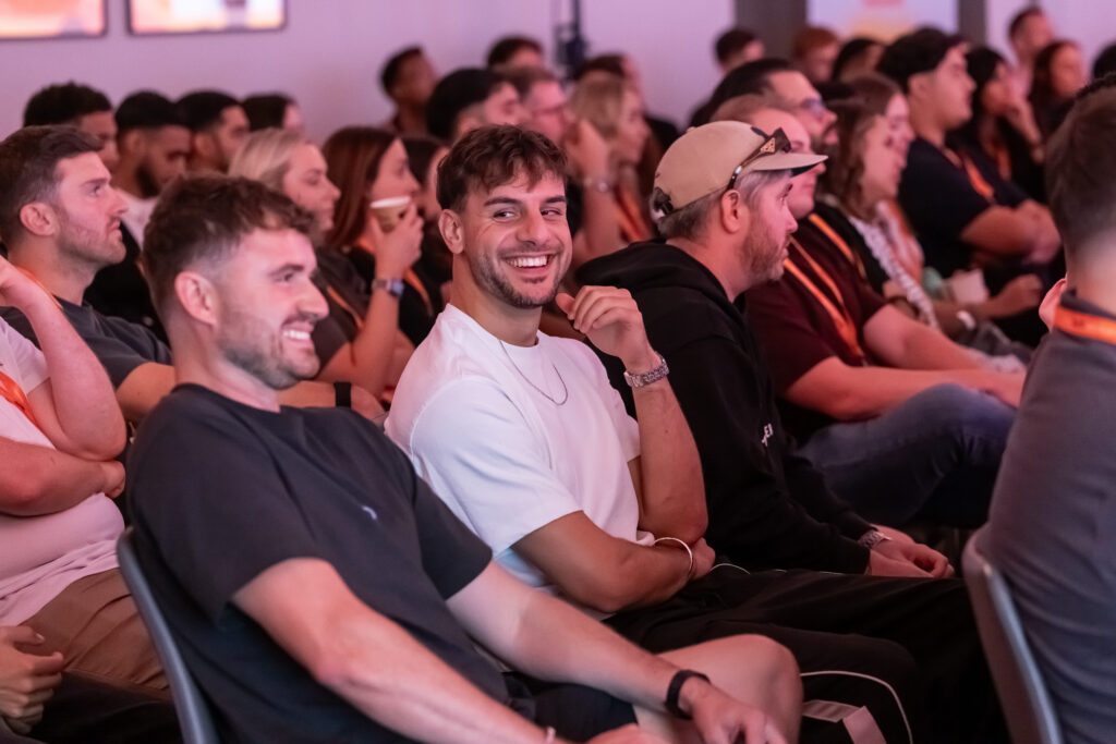 A smiling man in a white T-shirt sitting in an audience among other attendees at an advisory board event.