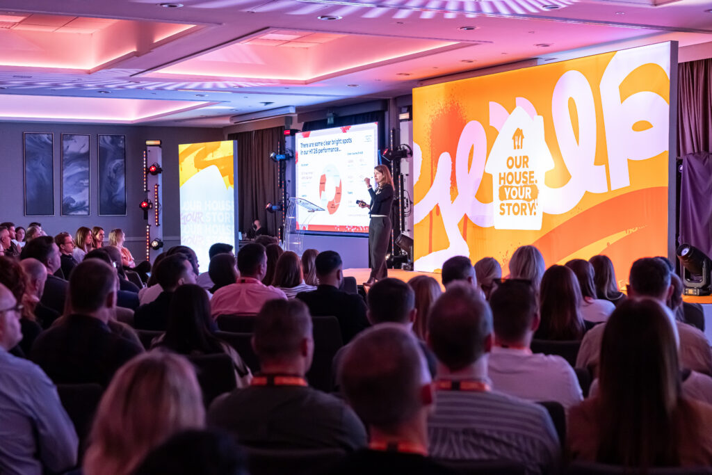 A speaker presents to an audience at an advisory board event in a conference hall with large screens displaying the text Our house. Your story. and corporate performance data.