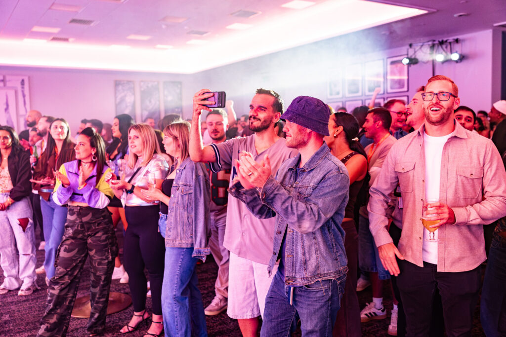 A diverse crowd gathers in a dimly lit, purple-hued advisory board event space, with some attendees applauding and others taking photos with their mobiles.