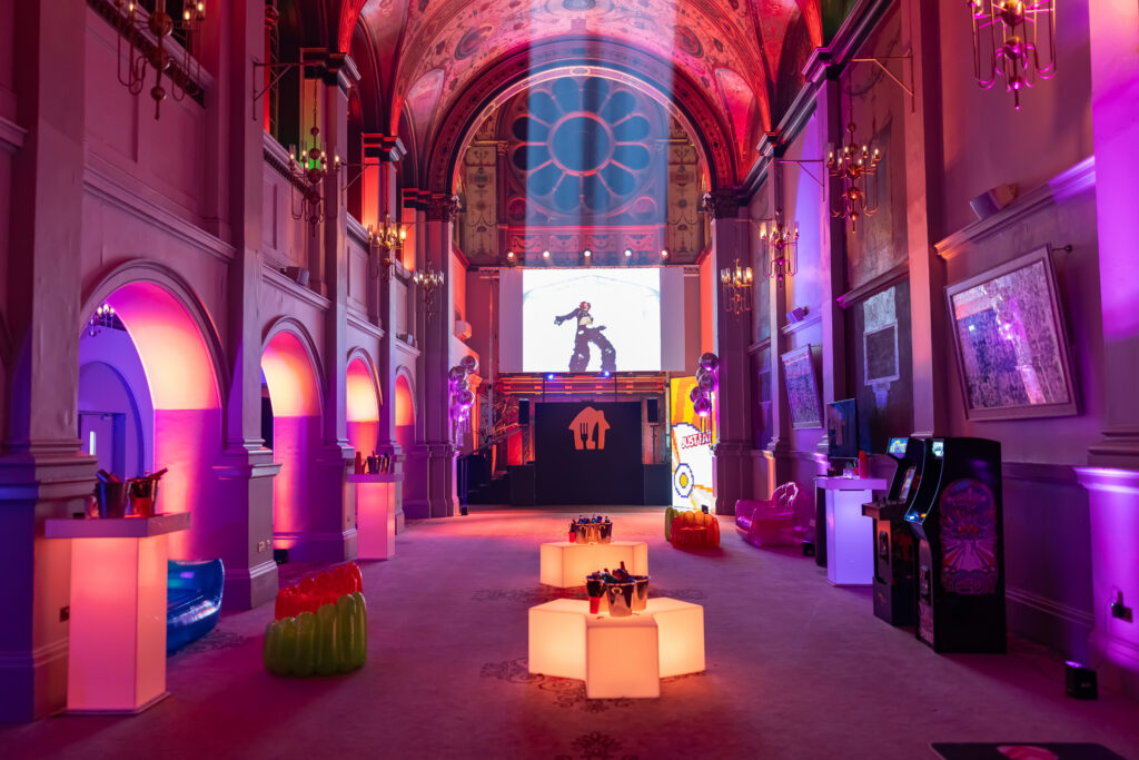 A large, pink-lit advisory board event hall with a projection screen displaying a performer, illuminated hexagonal furniture, and arcade games.