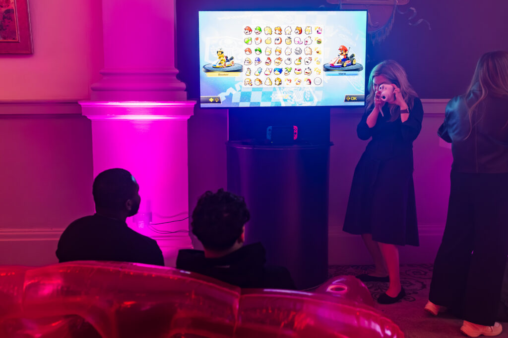 Two people sit in front of a screen displaying the Mario Kart 8 character selection menu at an advisory board event whilst a woman photographs the set-up.