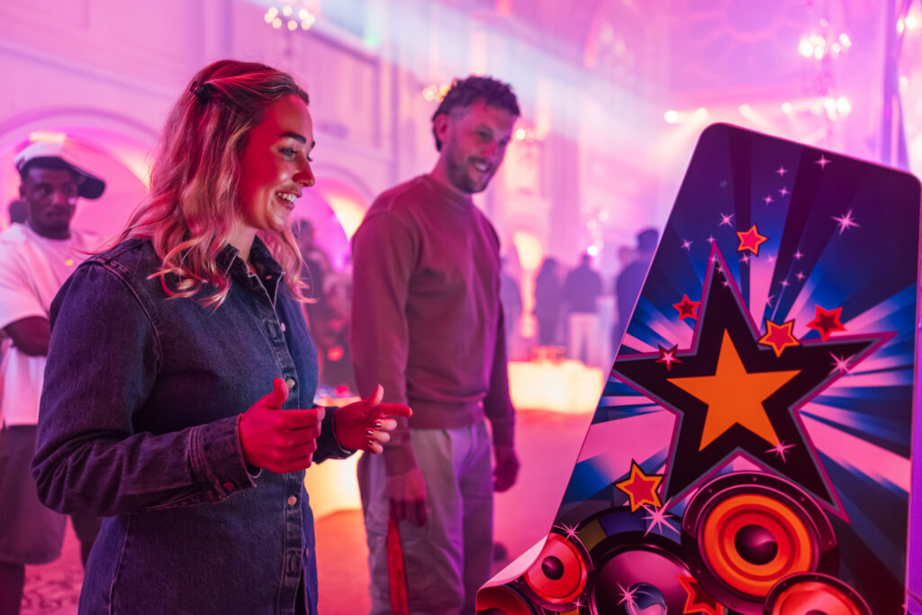 A woman smiles whilst playing an arcade game at an advisory board event in a room lit with pink light, as a man watches her.