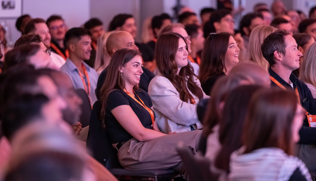 A diverse audience sits in an auditorium during a Sales Summit event, listening and smiling whilst watching a presentation.