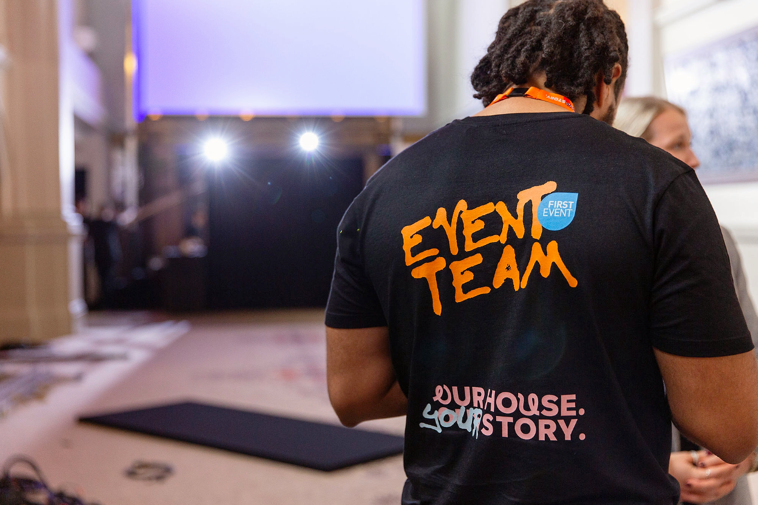 A person wears a black T-shirt for event engagement featuring EVENT TEAM in orange, OURHOUSE. YOURSTORY. in pink, and a blue FIRST EVENT circle logo.
