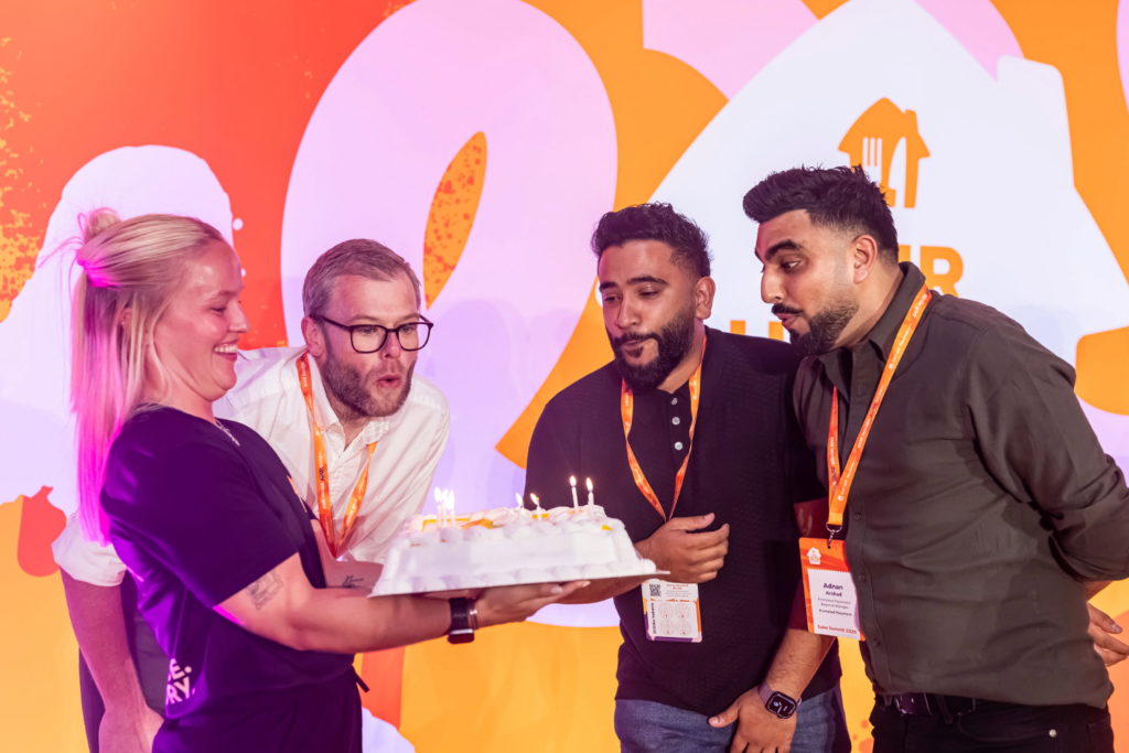 Four colleagues gather round a birthday cake with lit candles to build brand loyalty through events as one person blows them out.