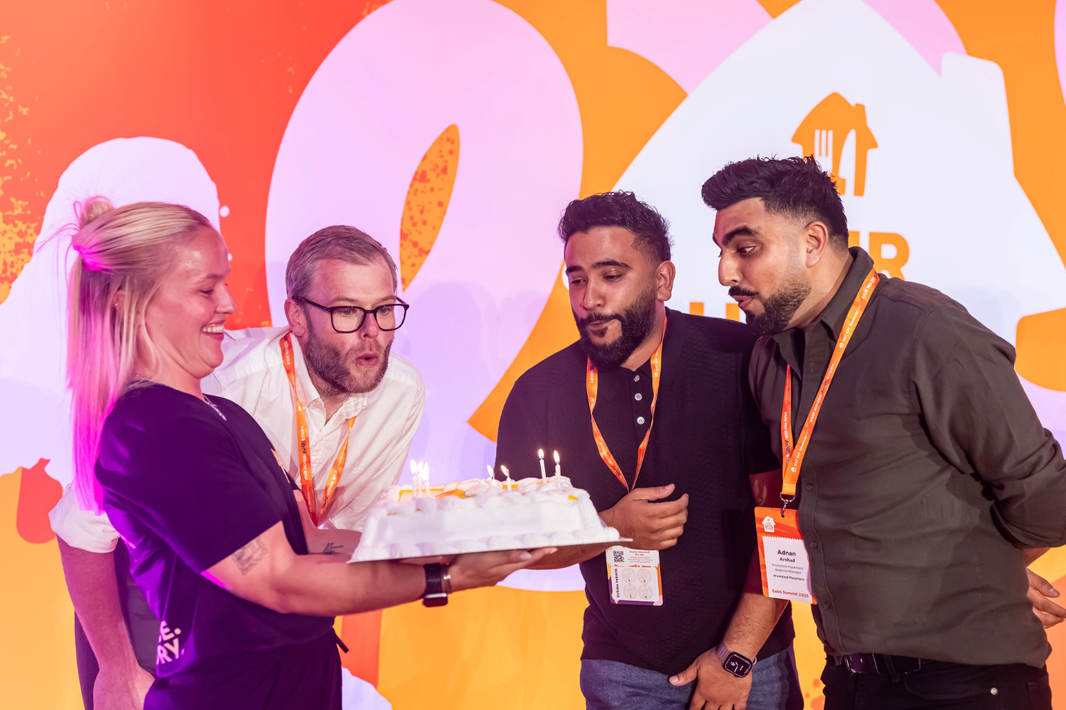 Four colleagues gather round a birthday cake with lit candles to build brand loyalty through events as one person blows them out.